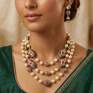 May include: A three-strand pearl necklace with gold accents and red gemstone embellishments. The necklace features ornate, round pendants and is paired with matching earrings. The jewelry is displayed against a green garment.