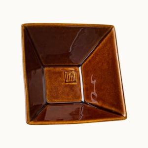 Puede incluir: Un plato cuadrado de cerámica marrón con acabado brillante. El plato tiene un diseño geométrico con lados angulados y un centro empotrado. Un pequeño emblema estilizado es visible en el centro. El plato es probablemente para servir o exhibir.