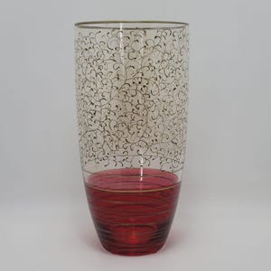 Könnte beinhalten: Eine hohe, transparente Glasvase mit rotem Boden und goldenen Akzenten. Der obere Teil weist ein zartes, wirbelndes Muster in Gold auf. Die zylindrische Vase eignet sich zum Präsentieren von Blumen oder als dekoratives Element. Höhe ca. 25 cm.