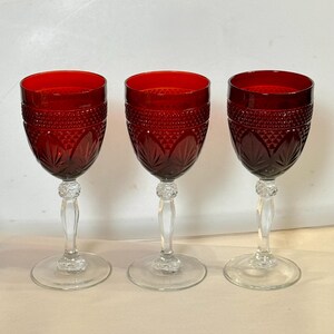 May include: Three ruby red wine glasses with clear stems and bases. The bowls have a diamond and leaf pattern. The stems have a decorative, textured design. The glasses are on a light background.