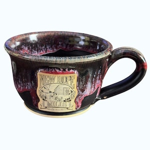May include: A ceramic mug with a dark, glossy finish, featuring a label that reads "NEW HOPE MILLS." The mug has a rounded shape with a handle and a contrasting red and black color scheme. The label includes an illustration of a building.