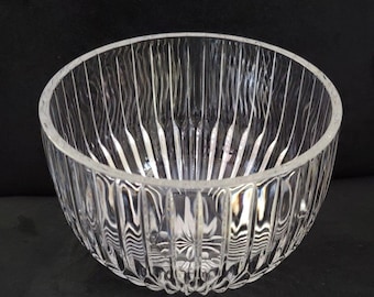 Könnte beinhalten: Eine klare Glasschale mit einem gerippten, vertikalen Muster. Die Schale hat eine abgerundete Form und steht auf einer dunklen Oberfläche. Das Glas ist transparent, so dass Licht hindurchscheinen kann, was das strukturierte Design hervorhebt. Diese dekorative Schale eignet sich zum Servieren oder Ausstellen.