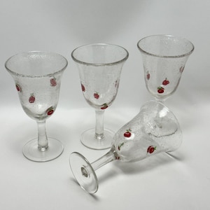 May include: Four clear wine glasses with a textured surface and a red strawberry design. The glasses have a rounded bowl, a long stem, and a circular base. One glass is lying on its side.