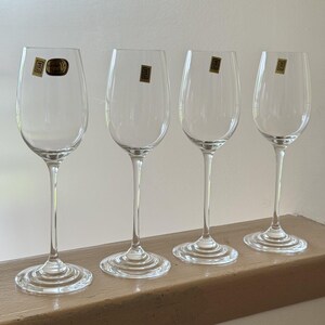 Op de afbeelding: Vier wijnglazen van helder glas met slanke stelen en ronde voeten. Elk glas is voorzien van een kleine rechthoekige goudkleurige en zwarte sticker met de letters LR en een kroonsymbool. Het eerste glas heeft ook een ronde gouden sticker.