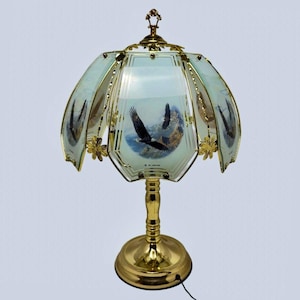 Puede incluir: Una lámpara de mesa con base de metal dorado pulido y pantalla de cristal de varios paneles. Cada panel de cristal presenta una ilustración impresa de águilas calvas volando sobre un paisaje montañoso. La pantalla está decorada con pequeños conectores metálicos dorados en forma de flor.