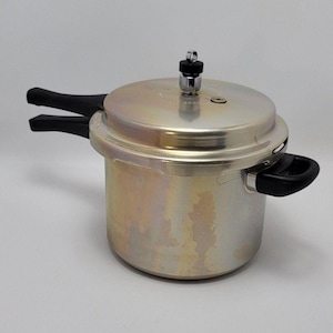 Puede incluir: Una olla a presión de metal plateado con cuerpo cilíndrico y tapa plana. La tapa cuenta con una válvula de presión vertical central y una pequeña ventilación circular. Dos asas de plástico negro sobresalen de los lados, con una asa colocada sobre la otra en el lado izquierdo.