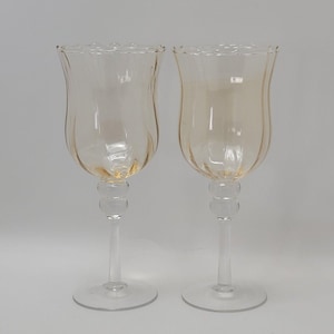 May include: Two elegant, amber-tinted wine glasses. Each glass features a tulip-shaped bowl with vertical ridges and a scalloped rim. The stem includes a decorative bulbous section, and the base is a clear, circular foot.