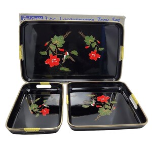May include: A black lacquerware tray set with three trays of varying sizes. Each tray features a floral design with red flowers, green leaves, and a hummingbird. The trays have gold trim and yellow woven handles. The box reads "Red Orchids 3 pc. Lacquerware Tray Set."