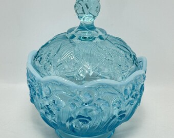 Könnte beinhalten: Eine hellblaue Glasschale mit passendem Deckel, die ein detailliertes, geprägtes Blumen- und Blattmuster auf der gesamten Oberfläche aufweist. Die Schale hat einen gewellten Rand, einen Standfuß und einen dekorativen Knauf auf dem Deckel.
