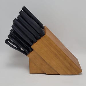 May include: A wooden knife block with a set of black-handled knives. The block is made of light brown wood and has a slanted design. The knives are neatly arranged, suggesting a kitchen utensil set.