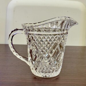 Op de afbeelding: Een heldere, vintage-stijl glazen creamer met een diamantgeslepen patroon. De creamer heeft een handvat en een tuit om gemakkelijk te schenken. Het glas is dik en heeft een getextureerd oppervlak.