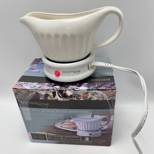 K&ouml;nnte beinhalten: Eine wei&szlig;e Keramik-Sauciere steht auf einer runden elektrischen W&auml;rmestation mit roter Kontrollleuchte. Die Basis ist mit Kitchen Living beschriftet und hat ein wei&szlig;es Stromkabel. Das Ger&auml;t steht auf der originalen Produktverpackung aus Karton.