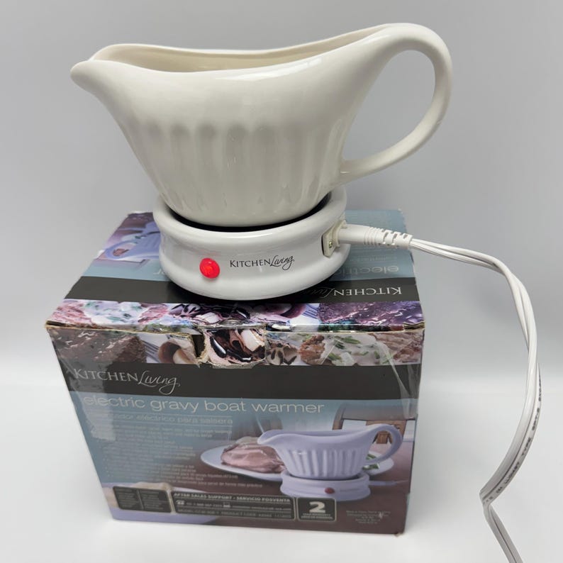 K&ouml;nnte beinhalten: Eine wei&szlig;e Keramik-Sauciere steht auf einer runden elektrischen W&auml;rmestation mit roter Kontrollleuchte. Die Basis ist mit Kitchen Living beschriftet und hat ein wei&szlig;es Stromkabel. Das Ger&auml;t steht auf der originalen Produktverpackung aus Karton.