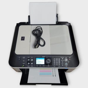 May include: A Canon MX350 all-in-one printer in a black and light gray finish. The device features a top paper tray with a sheet of paper, a coiled black power cord resting on the scanner lid, and a front output tray displaying a test print page.