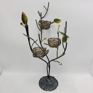 Op de afbeelding: Een decoratieve metalen boomsculptuur met drie geweven nestvormige kaarshouders. De boom heeft een donkere afwerking met groene en gele accenten, waaronder bladeren en kleine bolvormige details. De basis is een rond, getextureerd ontwerp.