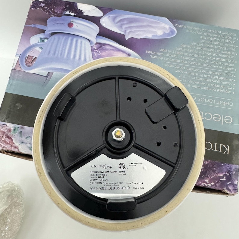 K&ouml;nnte beinhalten: Die Unterseite eines elektrischen So&szlig;enw&auml;rmers von Kitchen Living, Modell CCW-008-1, mit einem schwarzen runden Boden, drei schwarzen Standf&uuml;&szlig;en, einem zentralen Metallkontaktpunkt und einem silbernen Etikett mit elektrischen Angaben.