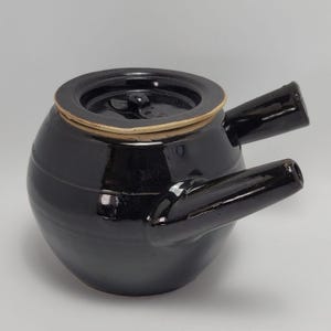 Könnte beinhalten: Eine schwarze Keramik-Teekanne mit Deckel und zwei Ausgüssen. Die Teekanne hat einen runden Körper und eine glänzende Oberfläche. Der Deckel ist dunkel mit einem hellen Rand. Die Ausgüsse sind abgewinkelt.