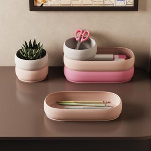 Könnte beinhalten: Eine Sammlung von Schreibtisch-Organisatoren in Rosa- und Weißtönen. Das Set umfasst eine kleine Sukkulente in einem zweifarbigen Topf, einen Stapel Tabletts und ein Tablett mit Stiften. Eine rosa Schere liegt in einem der Organizer.