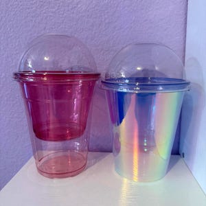 May include: Two plastic cups with dome lids. The cup on the left is transparent pink with a matching pink inner insert. The cup on the right features an iridescent, holographic finish that reflects multiple colors, paired with a clear dome lid.