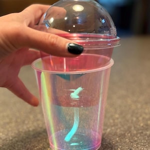 May include: A clear, iridescent plastic cup with a matching lid. The cup has a straw and a small, white design at the bottom. The cup is held by a hand with a black painted fingernail.