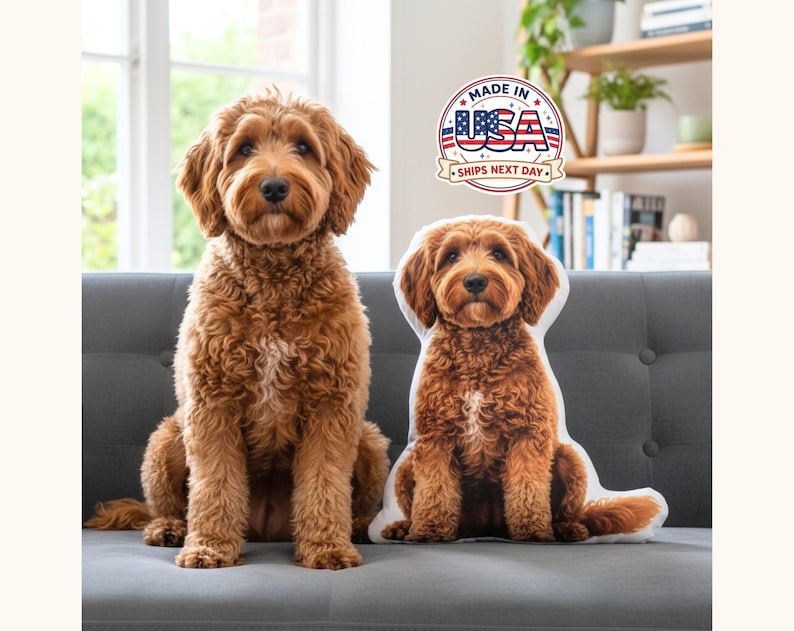 May include: A real-life golden doodle dog sits next to a dog-shaped pillow on a gray couch. The pillow is a replica of the dog. The image includes a "Made in USA" logo.
