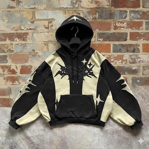 May include: Black and beige hoodie with a front pocket and a hood. The hoodie features star and abstract designs on the front, sleeves, and hood. The hoodie is hanging against a brick wall.
