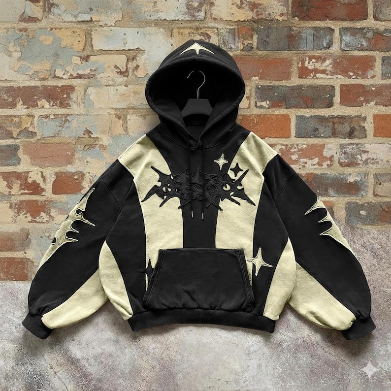 May include: Black and beige hoodie with a front pocket and a hood. The hoodie features star and abstract designs on the front, sleeves, and hood. The hoodie is hanging against a brick wall.