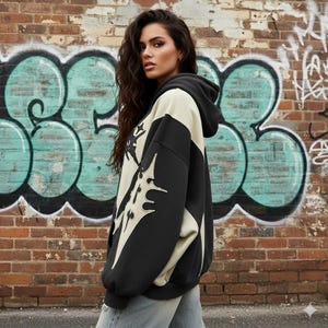 May include: Black and cream-colored hoodie with a unique design. The hoodie features a large graphic on the front and sleeves. The model is wearing light-wash jeans. The background is a brick wall with graffiti.