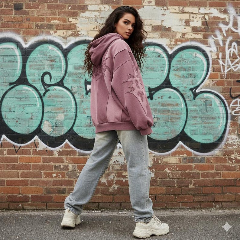 May include: A mauve hoodie with embroidered detailing on the back and sleeves, paired with light wash jeans and white sneakers. The background features a brick wall with teal graffiti art.