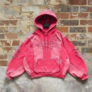 May include: A red hoodie with a front pocket and a hood. The hoodie features embroidered designs on the front, sleeves, and hood. The sleeves are long and the material appears to be a soft fabric.