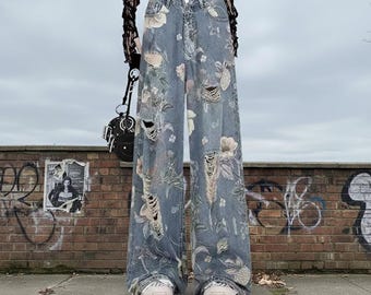May include: Light blue wide-leg jeans with floral print and distressed details. The jeans feature a high waist and are worn with white sneakers. The floral pattern includes pink, white, and green flowers.