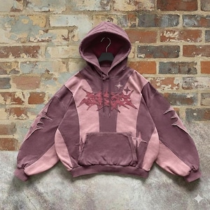 May include: A mauve and pink hoodie with a front pocket and a hood. The hoodie features a starburst design on the chest and sleeves, with star accents. The garment is displayed against a brick wall.