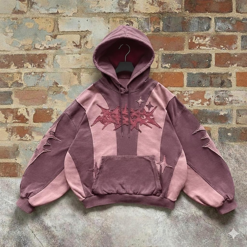 May include: A mauve and pink hoodie with a front pocket and a hood. The hoodie features a starburst design on the chest and sleeves, with star accents. The garment is displayed against a brick wall.