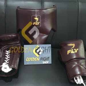 May include: A boxing set in brown leather, including gloves, headgear, and a protective cup. The gloves have white laces and the headgear has a yellow and black logo. The set is displayed on a black surface.