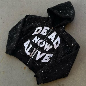 Pu&ograve; includere: Felpa nera con zip e testo bianco "DEAD NOW ALIVE". La felpa con cappuccio ha un cappuccio ed &egrave; adornata da piccoli abbellimenti scintillanti. La cerniera &egrave; argentata.