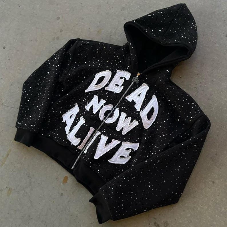 Pu&ograve; includere: Felpa nera con zip e testo bianco "DEAD NOW ALIVE". La felpa con cappuccio ha un cappuccio ed &egrave; adornata da piccoli abbellimenti scintillanti. La cerniera &egrave; argentata.