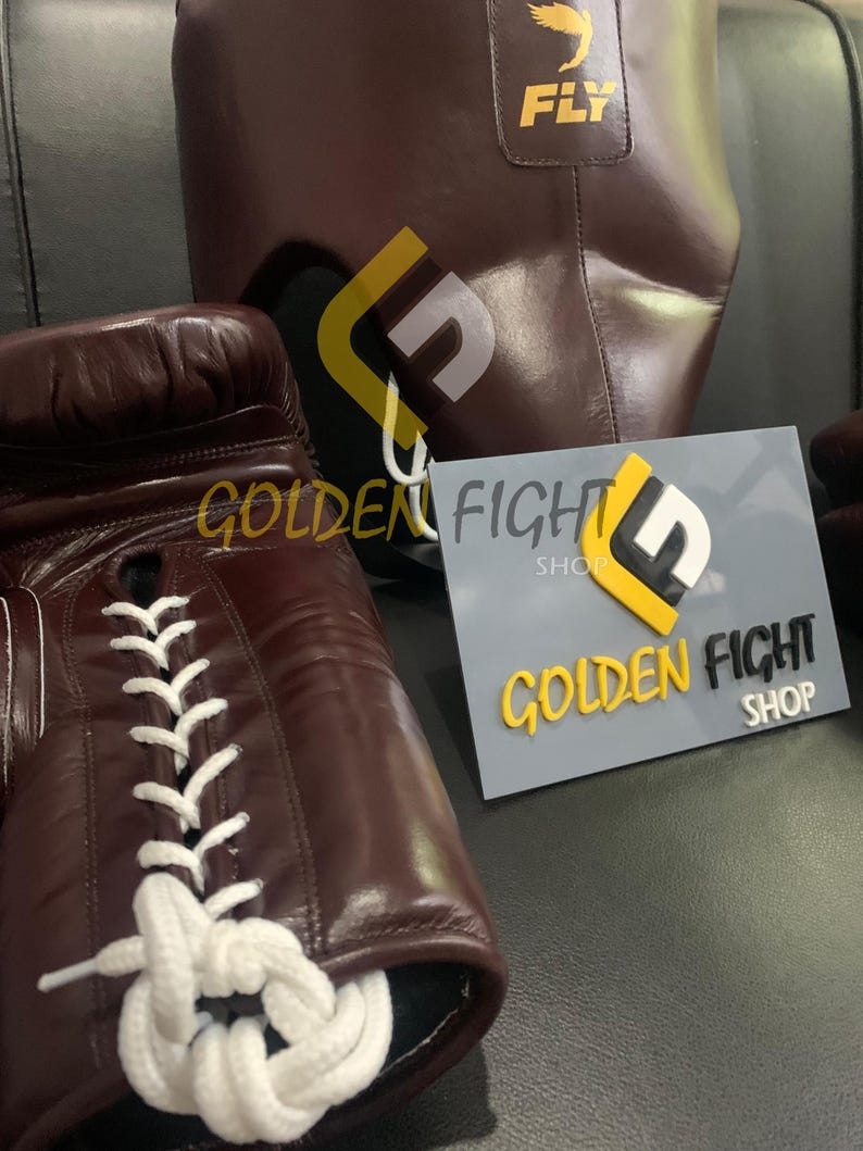 May include: Brown leather boxing gloves with white laces and a matching headgear. The headgear has the word "FLY" printed on it. A sign that says "GOLDEN FIGHT SHOP" is also in the image.