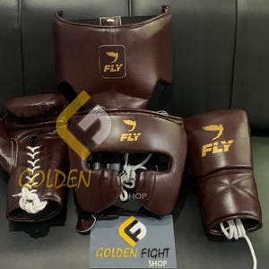 May include: A set of brown boxing gear, including a head guard, gloves, and a face protector. Each piece has the "FLY" logo in gold. The gear is displayed on a dark surface, with a "GOLDEN FIGHT SHOP" sign.