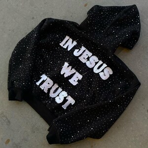 Pu&ograve; includere: Felpa con cappuccio nera con la scritta bianca "IN JESUS WE TRUST", impreziosita da numerosi piccoli accenti scintillanti. La felpa con cappuccio ha un cappuccio e un design classico, adatta all'abbigliamento casual.