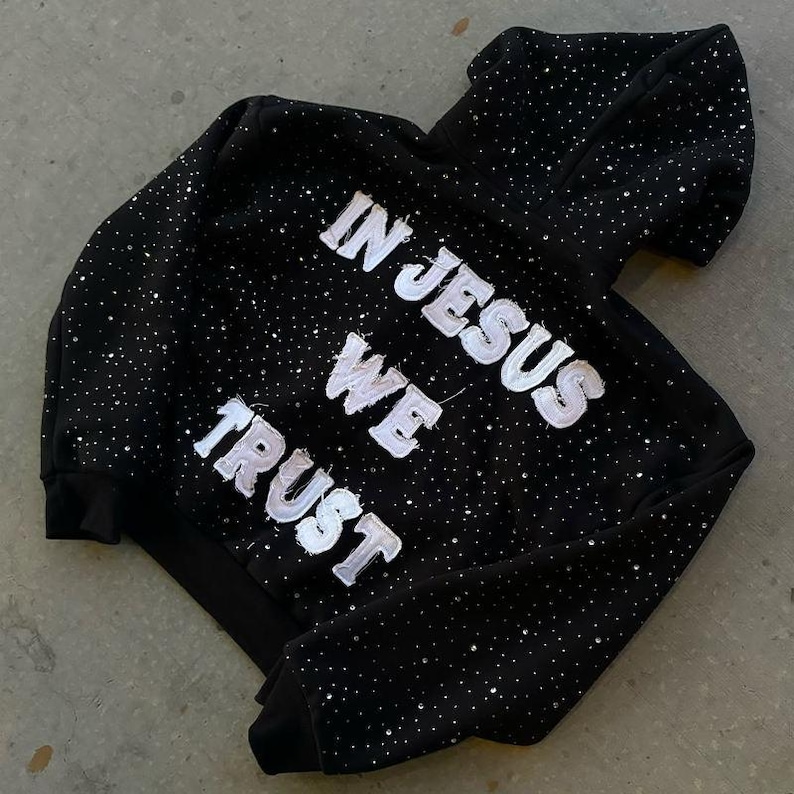 Pu&ograve; includere: Felpa con cappuccio nera con la scritta bianca "IN JESUS WE TRUST", impreziosita da numerosi piccoli accenti scintillanti. La felpa con cappuccio ha un cappuccio e un design classico, adatta all'abbigliamento casual.