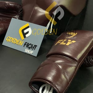 May include: A close-up of boxing equipment, including a pair of brown leather boxing gloves with white laces, a matching heavy bag, and a sign that reads "GOLDEN FIGHT SHOP" and "FLY". The gloves and bag have the "FLY" logo.