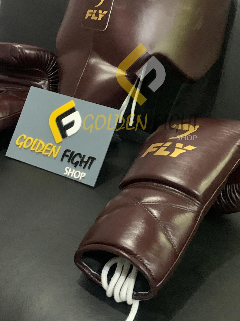 May include: A close-up of boxing equipment, including a pair of brown leather boxing gloves with white laces, a matching heavy bag, and a sign that reads "GOLDEN FIGHT SHOP" and "FLY". The gloves and bag have the "FLY" logo.