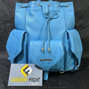 May include: A light blue leather backpack with multiple pockets and a drawstring closure. The backpack features a top handle, side pockets, and a front flap pocket. The text "GOLDEN FIGHT SHOP" is visible on a sign in front of the backpack.