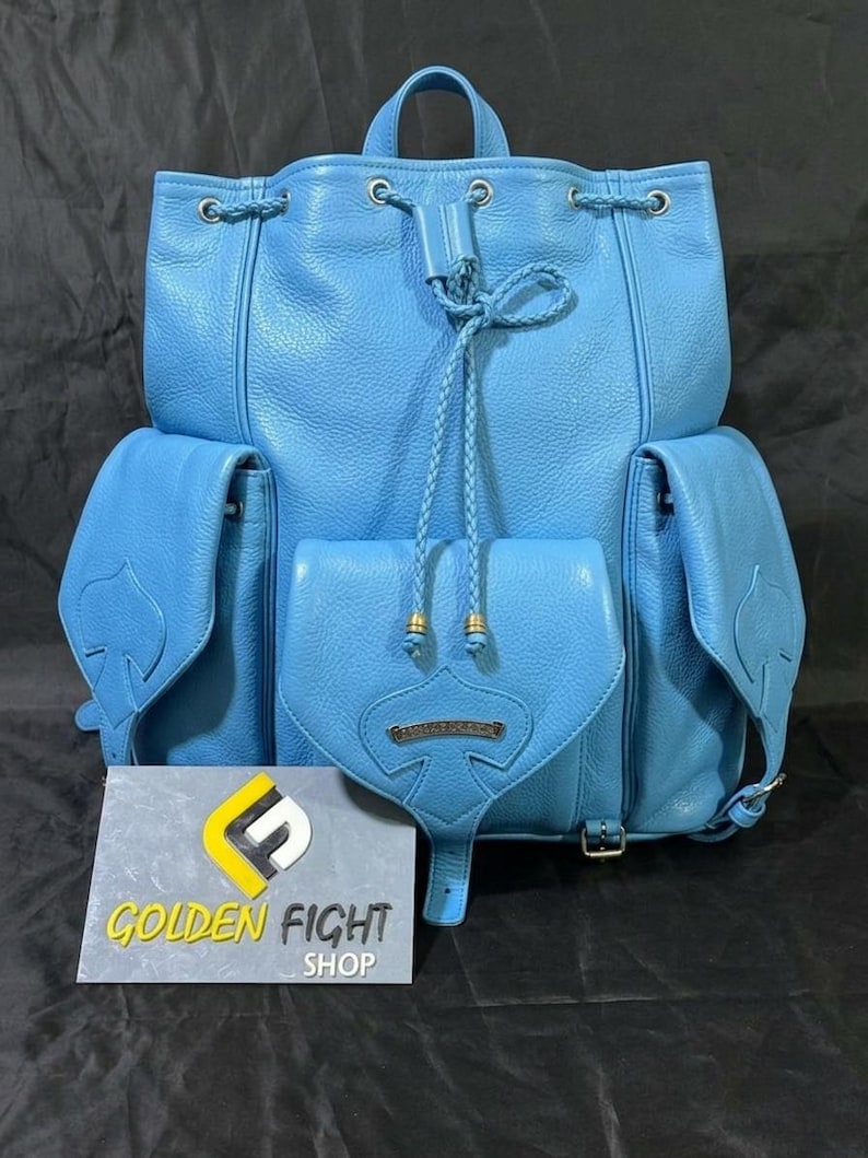 May include: A light blue leather backpack with multiple pockets and a drawstring closure. The backpack features a top handle, side pockets, and a front flap pocket. The text "GOLDEN FIGHT SHOP" is visible on a sign in front of the backpack.