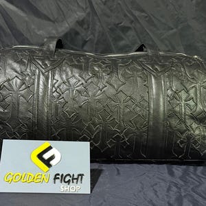 K&ouml;nnte beinhalten: Schwarze Leder-Duffel-Tasche mit sich wiederholendem Kreuzmuster. Die Tasche hat zwei Griffe und einen Rei&szlig;verschluss. Die Tasche liegt auf einer dunklen Oberfl&auml;che. Ein Schild mit der Aufschrift "GOLDEN FIGHT SHOP" befindet sich im Vordergrund.