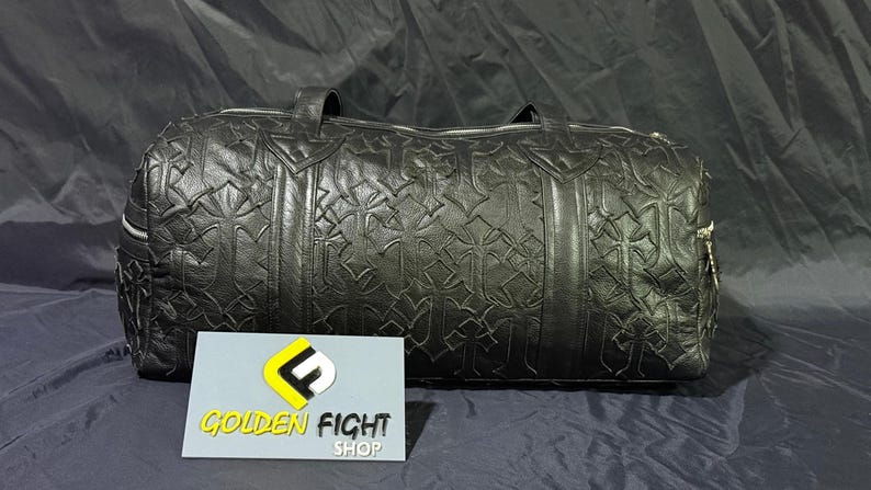 K&ouml;nnte beinhalten: Schwarze Leder-Duffel-Tasche mit sich wiederholendem Kreuzmuster. Die Tasche hat zwei Griffe und einen Rei&szlig;verschluss. Die Tasche liegt auf einer dunklen Oberfl&auml;che. Ein Schild mit der Aufschrift "GOLDEN FIGHT SHOP" befindet sich im Vordergrund.