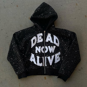 Pu&ograve; includere: Felpa nera con zip e cappuccio con la scritta bianca "DEAD NOW ALIVE" in lettere usurate. La felpa con cappuccio &egrave; ricoperta da piccoli abbellimenti scintillanti. &Egrave; un capo casual.