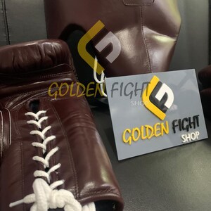 May include: Close-up of brown boxing gloves with white laces and a matching headgear. The headgear has the word "FLY" printed on it. A sign that says "GOLDEN FIGHT SHOP" is also visible.