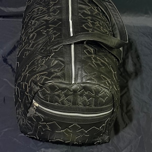 K&ouml;nnte beinhalten: Schwarze Reisetasche mit strukturiertem Muster und silbernen Rei&szlig;verschl&uuml;ssen. Die Tasche hat einen oberen Griff und einen Schultergurt. Auf der Tasche steht der Text "GOLDEN FIGHT SHOP" auf einem Schild.