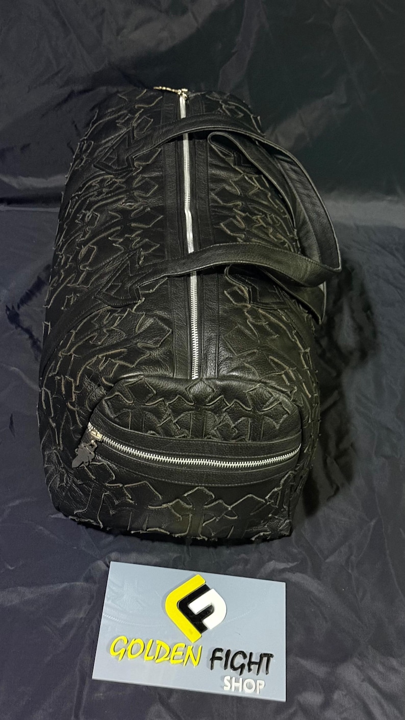 K&ouml;nnte beinhalten: Schwarze Reisetasche mit strukturiertem Muster und silbernen Rei&szlig;verschl&uuml;ssen. Die Tasche hat einen oberen Griff und einen Schultergurt. Auf der Tasche steht der Text "GOLDEN FIGHT SHOP" auf einem Schild.